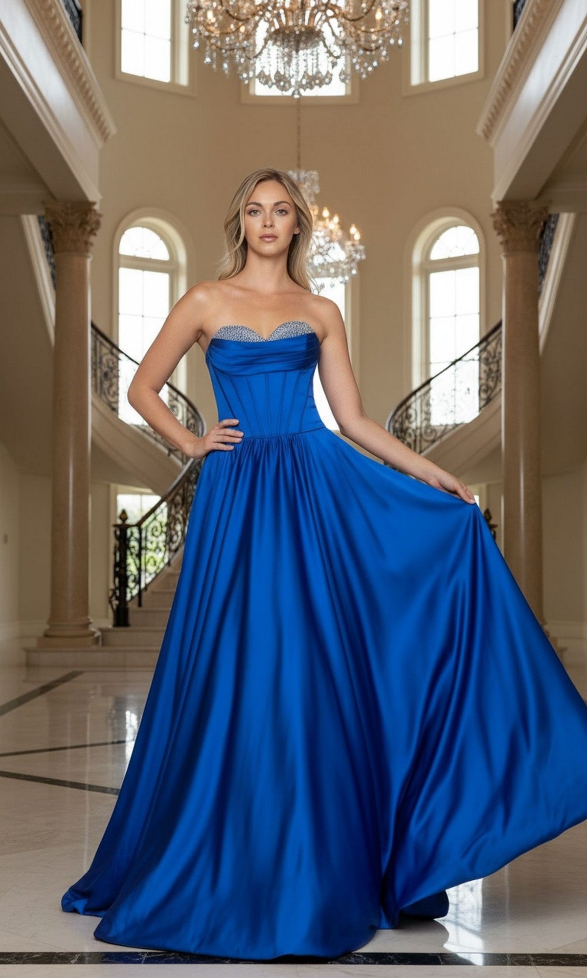 A woman stands in an elegant foyer wearing the Long Prom Dress: Poly USA 25136 in blue, a strapless gown with a flowing A-line silhouette, holding out one side of the dress.